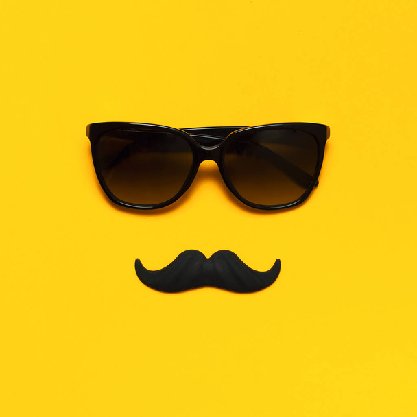 Creative party decoration concept. Black mustache, sunglasses, props for photo booths, carnival parties on yellow background top view flat lay copy space. Father's day Men's health awareness month - Fotó, kép