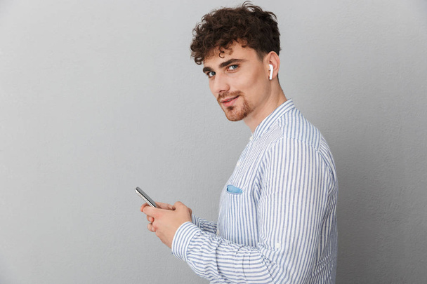 Image of unshaven young man with earpods looking at camera and holding cellphone - Фото, зображення