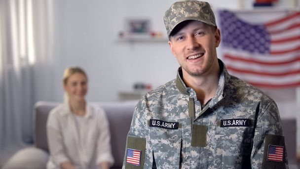 Sonriente militar estadounidense mirando a la cámara, esposa sentada en el fondo, apoyo
 - Foto, Imagen