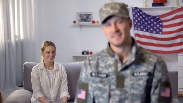 American soldier smiling at camera, proud wife sitting on sofa against US flag - Valokuva, kuva