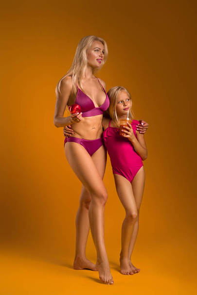 Блондинка мама держит яблоко и ее дочь в купальнике пьет свежий сок
. - Фото, изображение