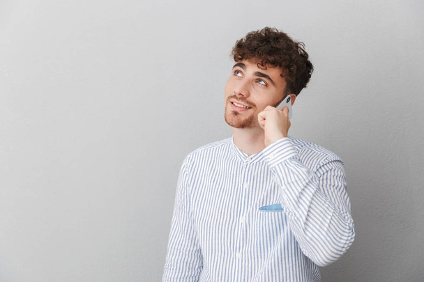 Portrait of brunette beautiful man dressed in shirt smiling while holding and talking on smartphone - Φωτογραφία, εικόνα
