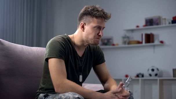 Depressed soldier with vodka bottle sitting on sofa, alcohol addiction, dementia - Фото, изображение