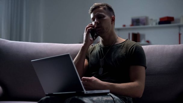 Male military spy sitting with laptop on sofa and talking smartphone at night - 写真・画像