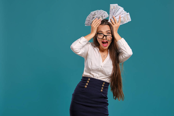 Chica morena en gafas, usando una falda corta negro y blusa blanca está posando sosteniendo un ventilador de billetes de cien dólares sobre un fondo azul
. - Foto, Imagen