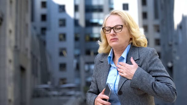 Aged female manager feeling chest pain holding phone, work nervousness, stress - Фото, изображение
