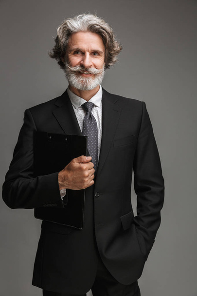 Image of happy adult businessman wearing formal black suit looking at camera and holding clipboard - Foto, afbeelding