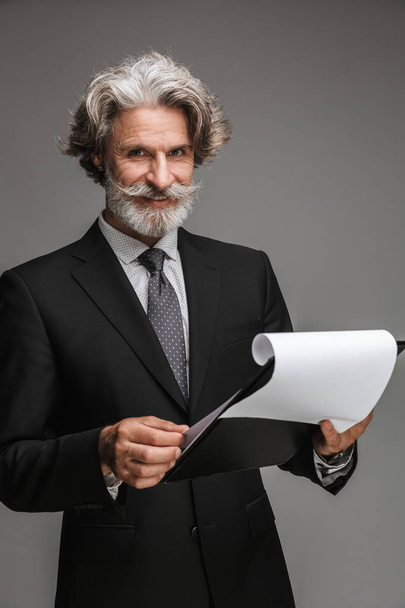Image of handsome adult businessman wearing formal black suit smiling and holding paper charts - Foto, afbeelding