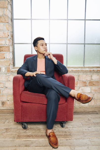 Ritratto di bell'uomo d'affari vietnamita alla moda che pensa a un nuovo progetto di start-up quando è seduto in poltrona di velluto rosso nell'ufficio del loft
 - Foto, immagini
