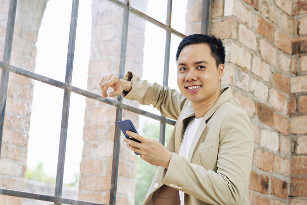 Портрет улыбающегося молодого азиатского бизнесмена, прислонившегося к большому офисному окну, СМС и улыбающегося перед камерой
 - Фото, изображение