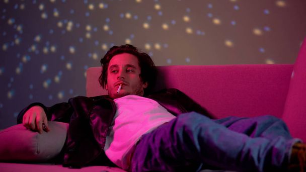 Relaxed young man smoking cigarette on couch, partying in club, relaxing - Photo, Image