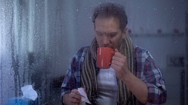 Ill male in scarf holding handkerchief and hot tea, treating cold, rainy weather - Foto, Imagem