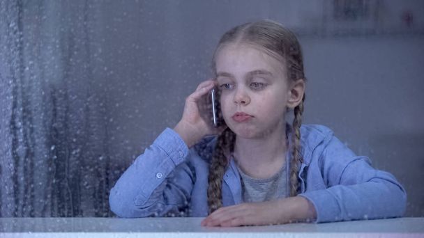 Sad little girl talking phone on rainy day, staying home alone, missing mother - Foto, imagen