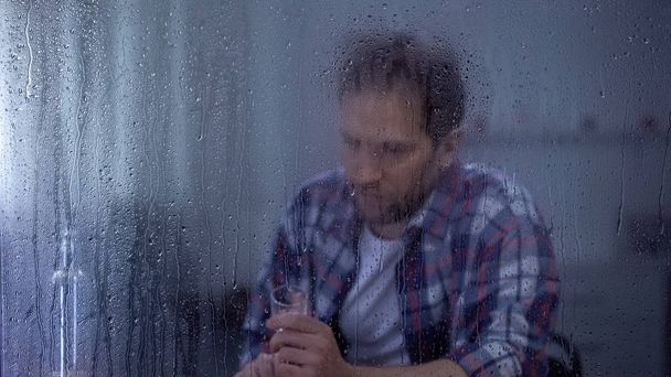Drunk man with glass of vodka behind rainy window, alcohol addiction problem - Valokuva, kuva