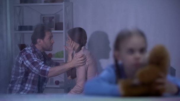 Sad girl with teddy bear on rainy day, father slapping mother on background - Photo, image