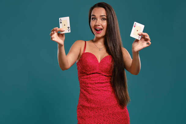 Brunette girl with a long hair, wearing a sexy red dress is posing holding two playing cards in her hands, blue background. - Foto, imagen