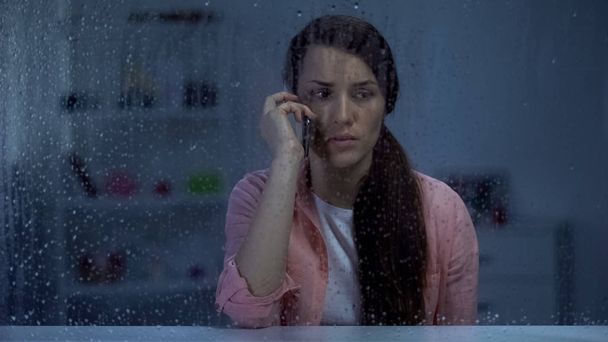 Sad woman talking phone behind rainy window, shocked by bad news from family - Photo, Image