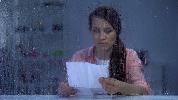 Depressed female reading letter behind rainy window, bad news from relatives - Photo, Image