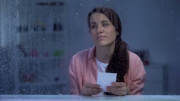 Sad woman with photo feeling sad behind rainy window, remembering first love - Photo, Image