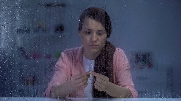 Sad woman holding engagement ring behind rainy window, upset after break up - Fotó, kép