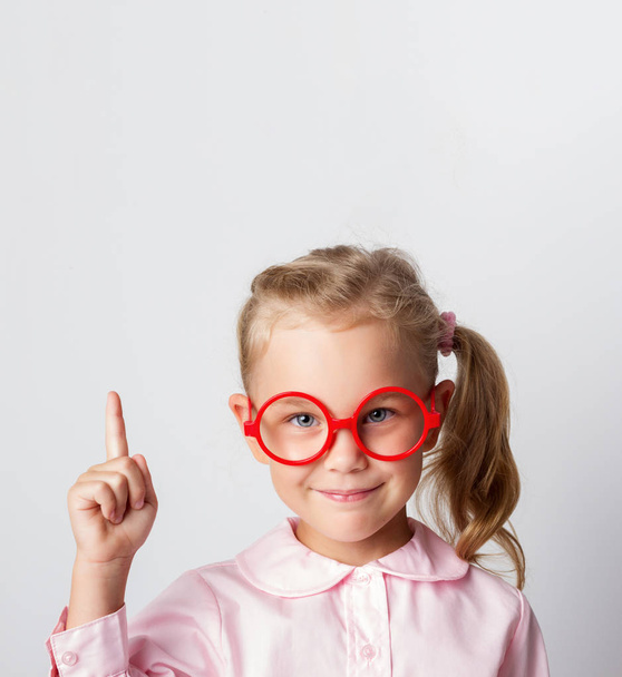 Portrait en gros plan d'une jolie écolière en lunettes rouges et un chemisier lumineux, pointant du doigt vers le haut, regardant la caméra avec un sourire, isolé sur un fond clair
 - Photo, image