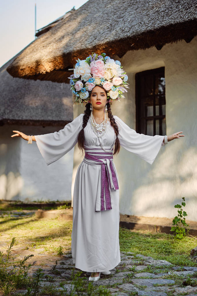 Brunette girl in a white ukrainian authentic national costume and a wreath of flowers is posing against a white hut. - Photo, Image