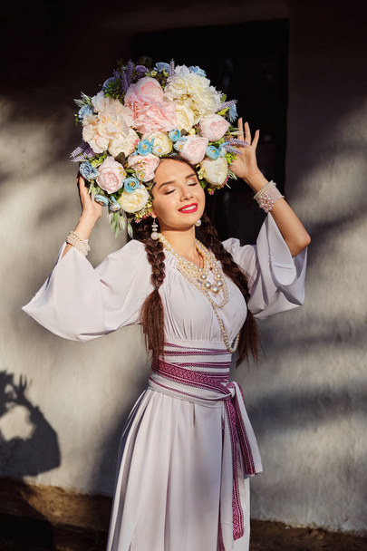 Brunette meisje in een wit Oekraïens authentiek nationaal kostuum en een krans van bloemen is poseren tegen een witte hut. Close-up. - Foto, afbeelding