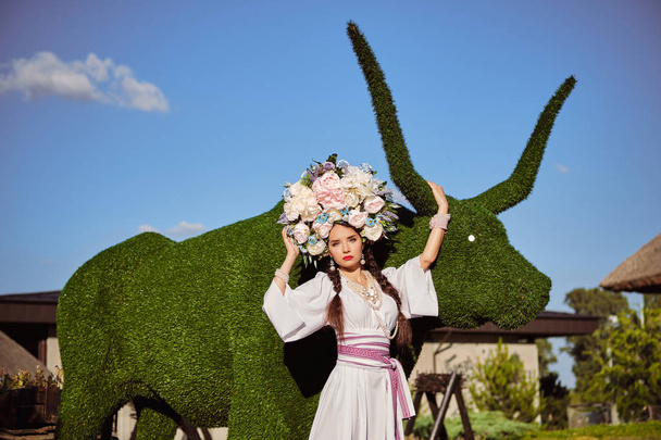 Дівчина брюнетка в білому українському автентичному національному костюмі і вінок з квітів позує проти зеленої бика різьблені з куща. - Фото, зображення