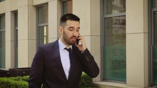 Young businessman feeling annoyed while talking on the cell phone outdoors near office building center. Man angry and sad. - Video, Çekim