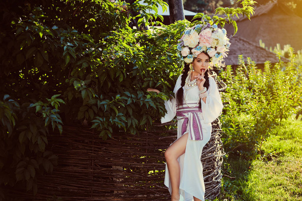Brunetka dziewczyna w białym ukraińskim autentycznym Narodowym stroju i Wieniec kwiatów pozuje się z drewnianej ogrodzenia wikliny. - Zdjęcie, obraz