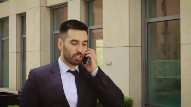 Young businessman feeling annoyed while talking on the cell phone outdoors near office building center. Man angry and sad. - Séquence, vidéo
