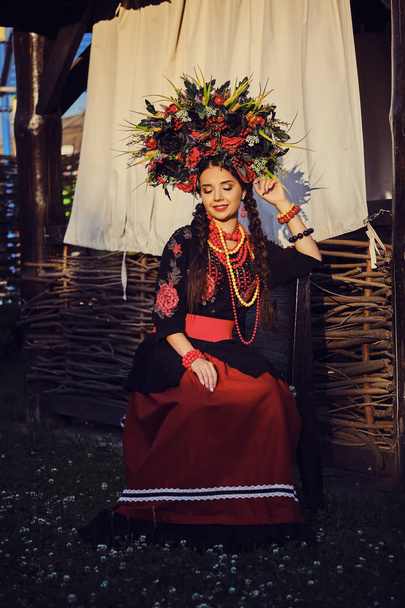 Μελαχρινή κορίτσι σε ένα λευκό ουκρανικό αυθεντικό εθνικό κοστούμι και ένα στεφάνι από λουλούδια ποζάρει σε μια βεράντα. Γκρο πλαν. - Φωτογραφία, εικόνα