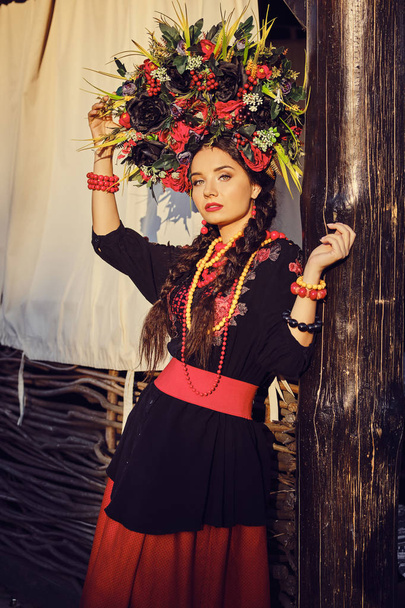Brunette meisje in een wit Oekraïens authentiek nationaal kostuum en een krans van bloemen poseren tegen een terras. - Foto, afbeelding