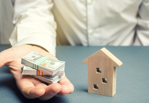 Businessman holds dollars in hands near wooden house. Real estate investment concept. Mortgage. Loan for housing. Credit. Rent. Rental. Buying a property. The accumulation of money. Selling a house - Photo, image