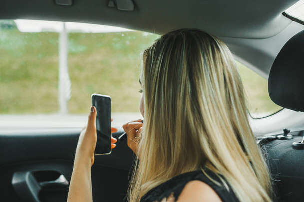 Ξανθιά γυναίκα κάθεται στο αυτοκίνητο με κινητό και καλλυντικά. Πίσω όψη. - Φωτογραφία, εικόνα