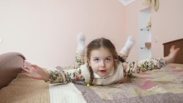 Little girl has fun at the bed at home. Cute child is relaxing and moves along the bed. Pretty girl is boring and plays with herself. - Video