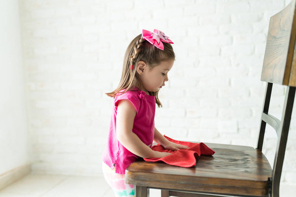 Seitenansicht des charmanten kleinen Mädchens, das zu Hause Holzstuhl putzt - Foto, Bild
