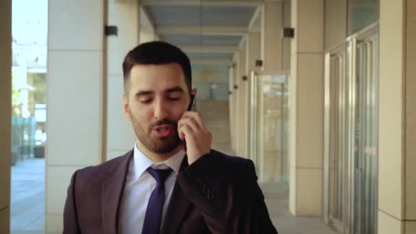 Close up view of businessman dressed in shirt talking smartphone while walks on the background of a modern building - Imágenes, Vídeo