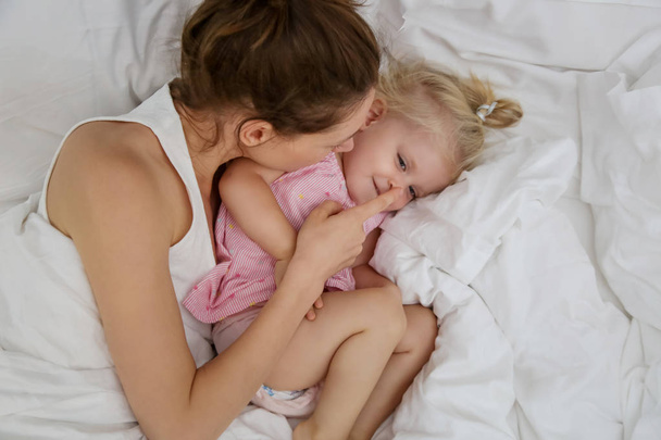 Mother and her infant baby cuddling in the bed, adorable blonde baby and her mom having fun, happy family life concept
 - Фото, изображение