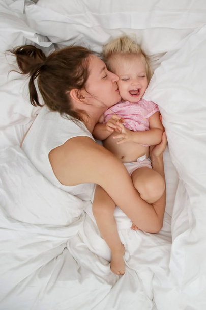 Mother and her infant baby cuddling in the bed, adorable blonde baby and her mum having fun, happy family life concept - Photo, Image