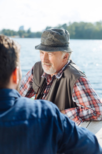 Bearded retired man speaking with son while sitting near lake - Foto, afbeelding