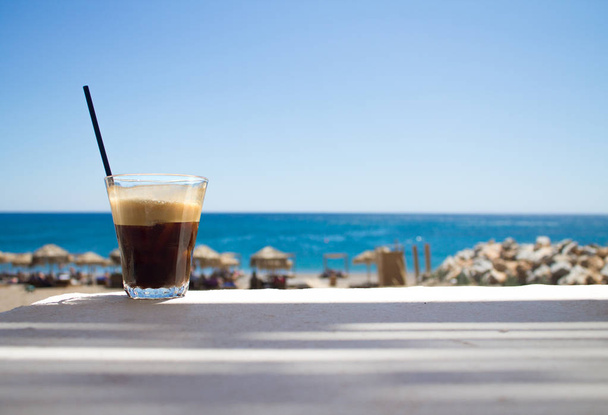 vaso con café helado fredo espresso o frappe en la esquina de la mesa con playa sobre fondo
 - Foto, imagen