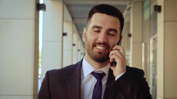 Close up view of smiling businessman dressed in shirt talking smartphone while walks on the background of a modern building - Filmagem, Vídeo