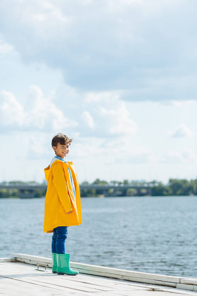 Handsome boy wearing bright rain boots standing near river - Φωτογραφία, εικόνα