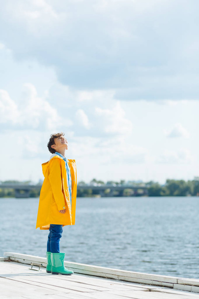 Cheerful boy wearing yellow raincoat looking at cloudy sky - Fotografie, Obrázek