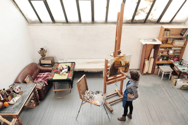 Bright art studio with a large window. Easels and canvases. Woman artist paints a picture on canvas. - Φωτογραφία, εικόνα