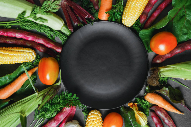 Creative layout made of corn, tomatoes, zucchini, carrot, beetroot, garlic, bean and black plate on dark background. Food concept. Flat lay, top view. Free space for your text. - Zdjęcie, obraz