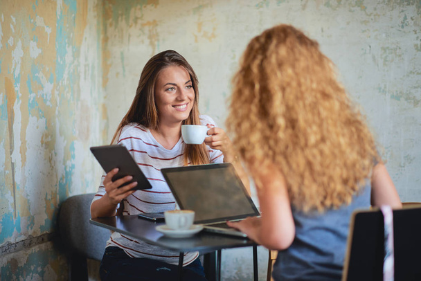 Дві жінки використовують технології і п'ють каву, сидячи в кафе
. - Фото, зображення