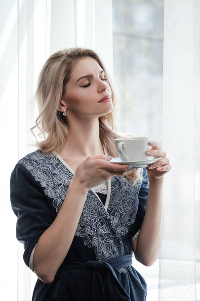 Beautiful young blonde woman in a blue robe by the window. Drinks coffee or tea from a white cup with a saucer. Morning, sunshine, bedroom window. - Photo, image