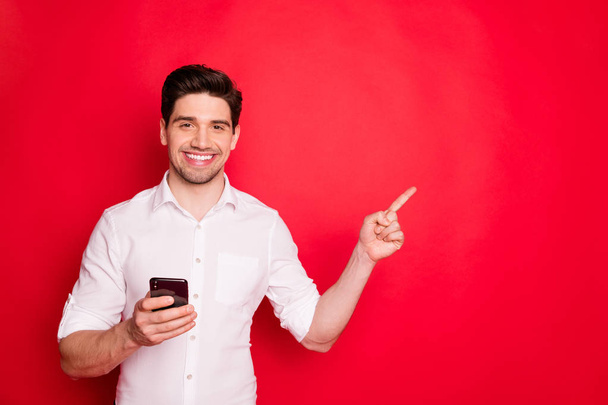 Photo of brunet man pointing at something near him holding phone with hand while isolated with red background - Fotografie, Obrázek